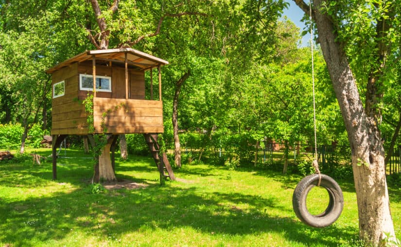 Tree house in a very green garden with a swing hanging in front of it